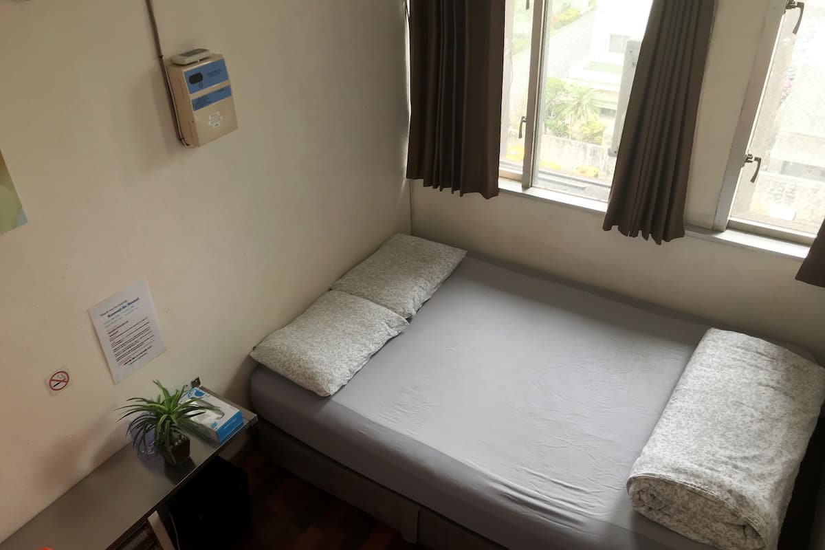 Una habitación pequeña con cama individual, ventana, mesita de noche con planta y un dispositivo en la pared.