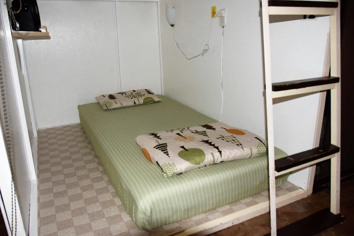 Una habitación pequeña con una cama individual, piso a cuadros, colcha a rayas verdes y almohada estampada.