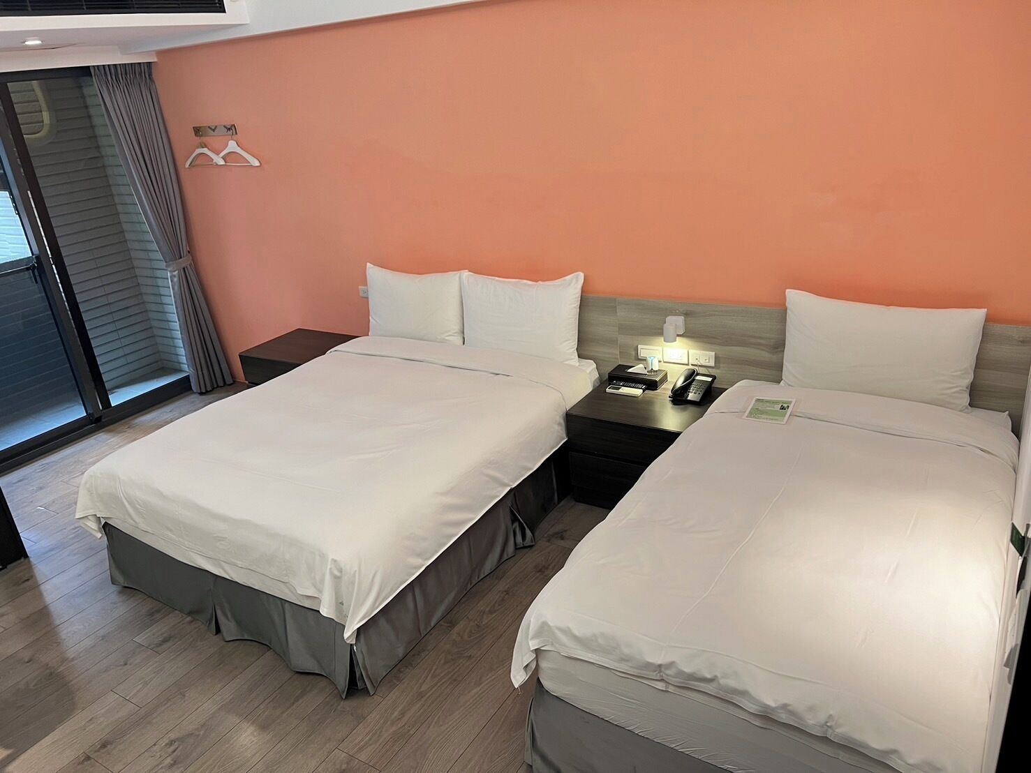 A hotel room with two beds, a nightstand with a phone and lamp, and a window with curtains.