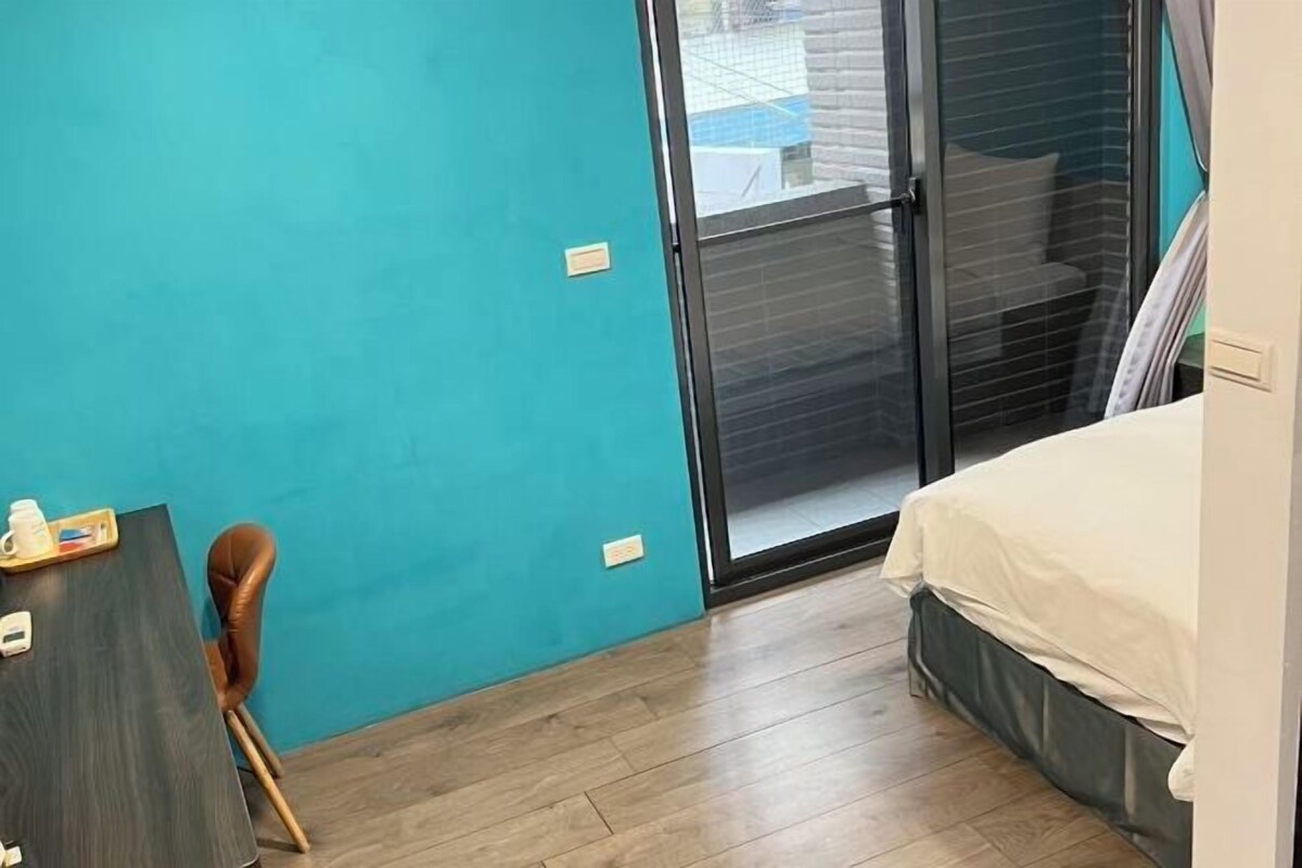 Una habitación pequeña con paredes verde azulado, una cama con edredón blanco, un escritorio con silla y un vidrio corredero que da a un balcón.
