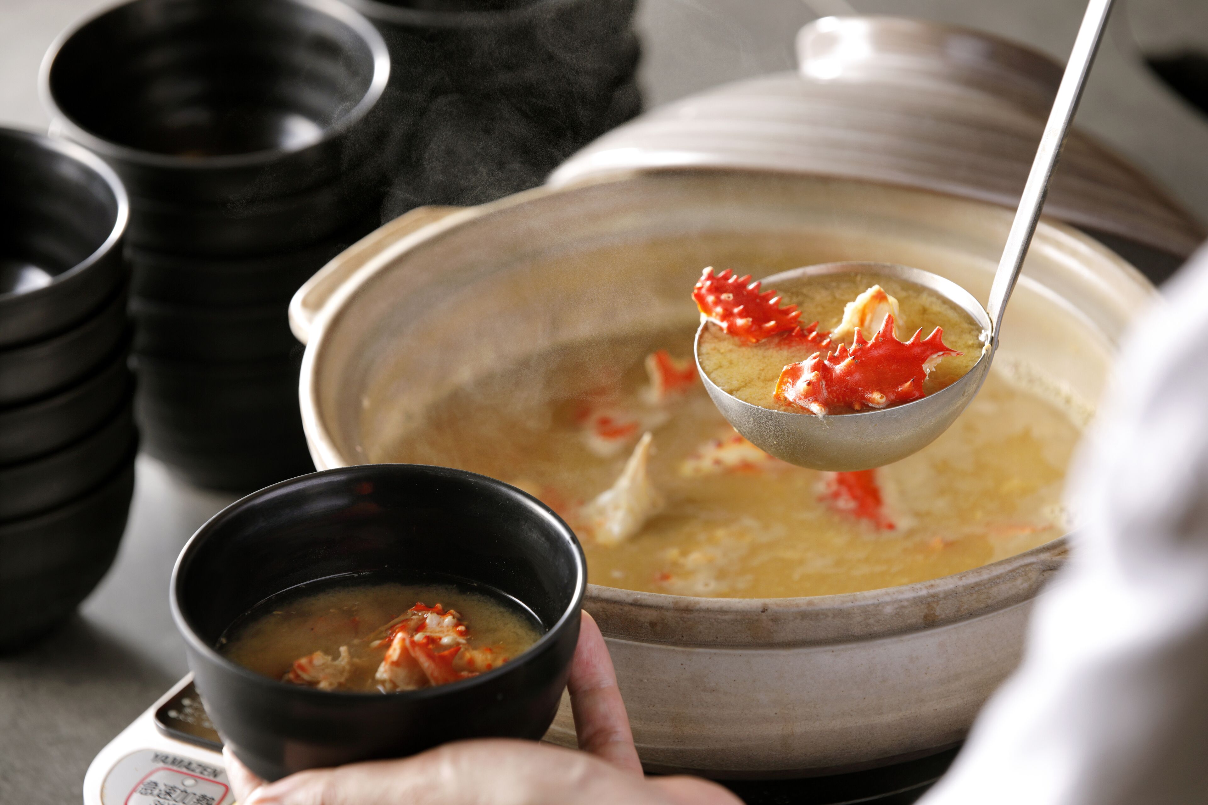A person holding a bowl of soup with a ladle, with a large pot of soup in the background.