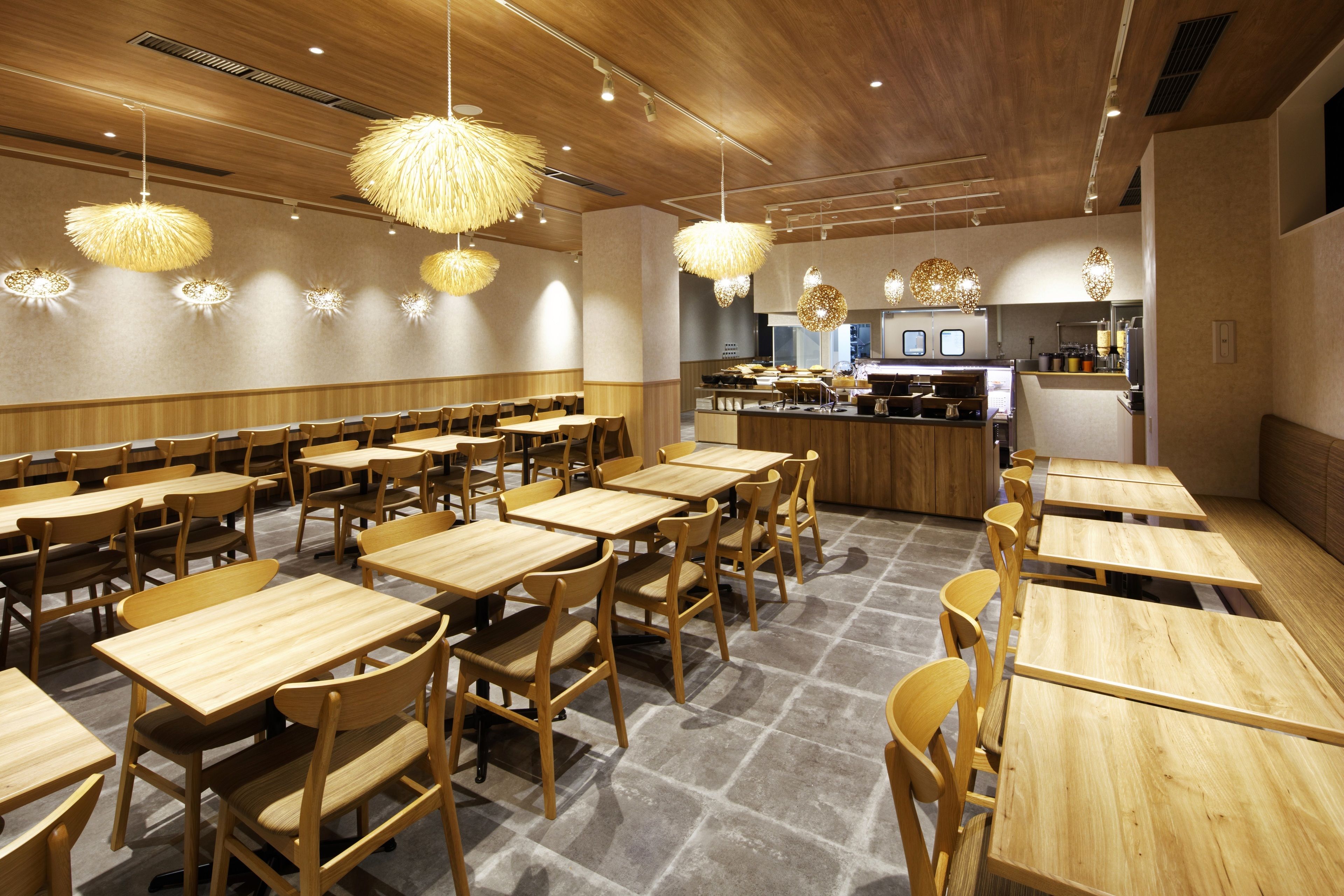 A restaurant with wooden tables and chairs, hanging light fixtures, and a kitchen area in the background.