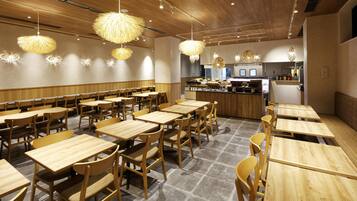 A restaurant with wooden tables and chairs, hanging light fixtures, and a kitchen area in the background.