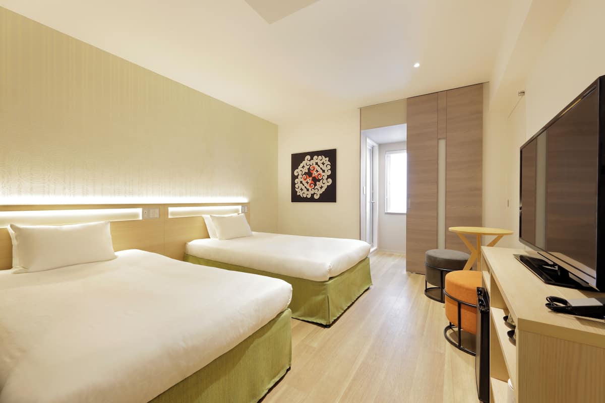 A hotel room with two beds, a desk, a television, and a small stool.