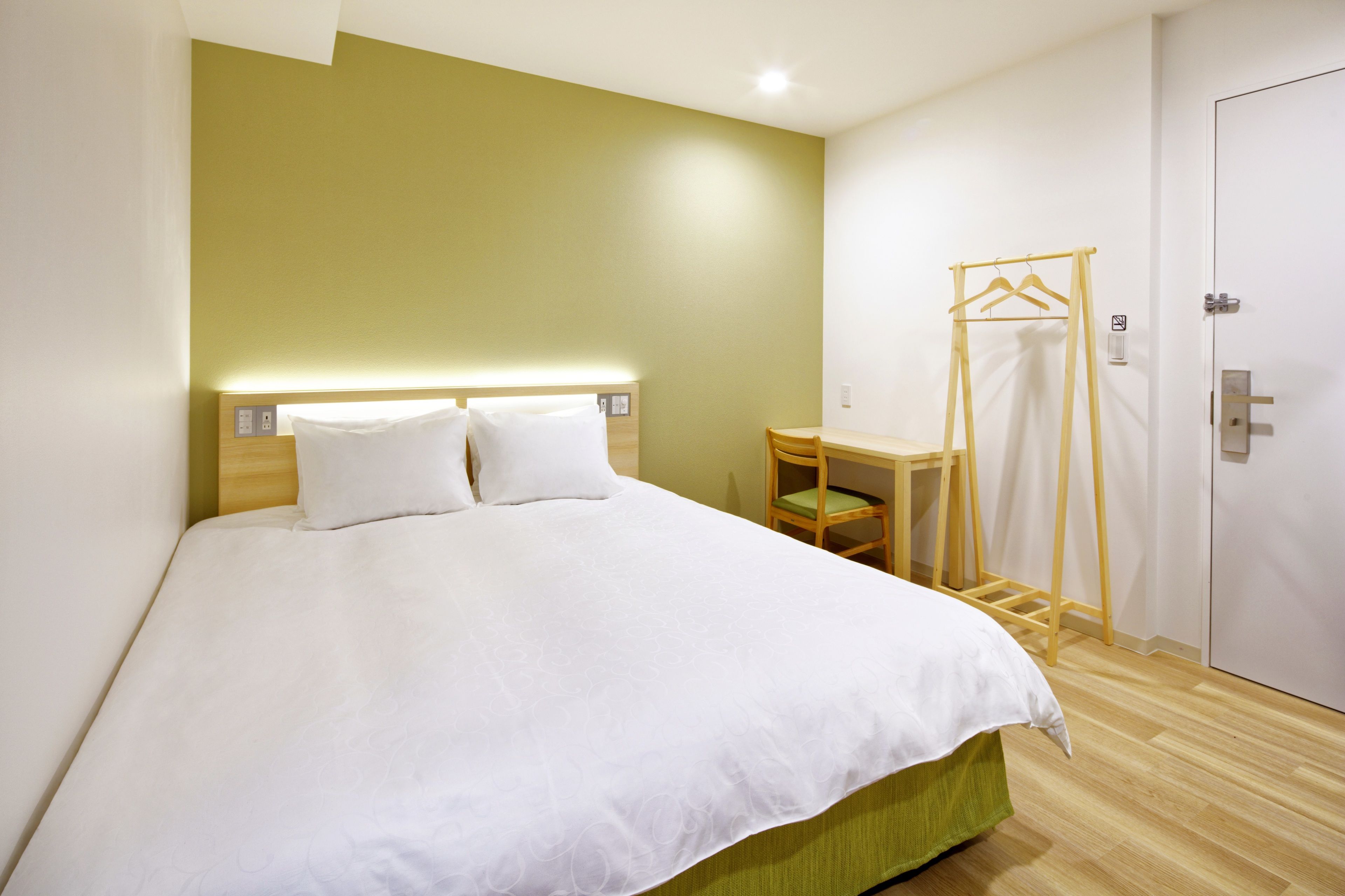 A neatly made bed with a white duvet, a bedside table with a chair, and a wooden hanger rack against a wall with a door.