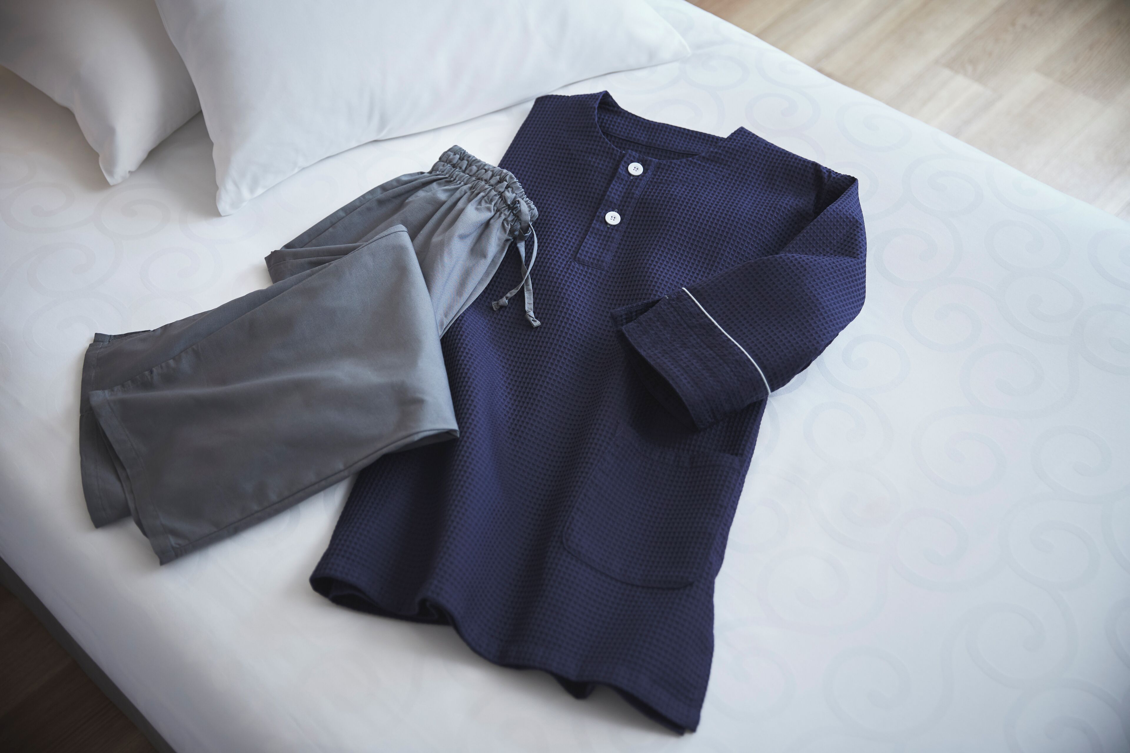 A neatly made bed with a white pillow and a blue and grey garment laid out on it.