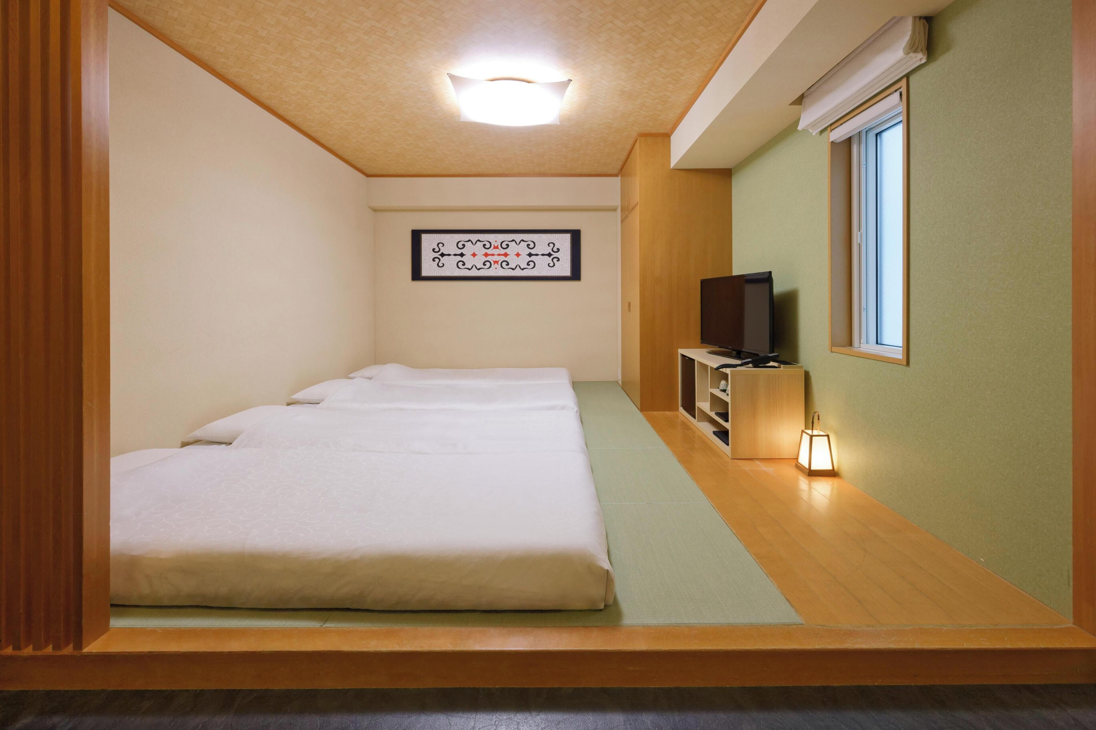A room with a bed, a television, a small table with a lamp, and a window.