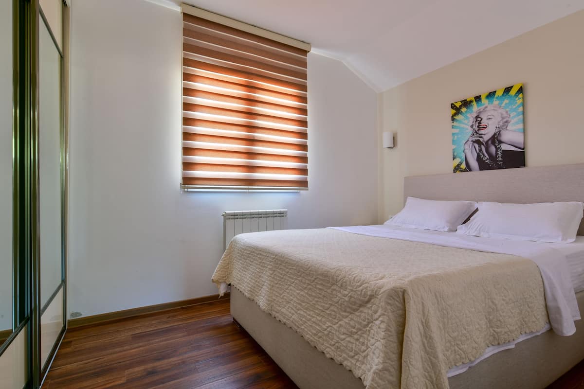 A bedroom with a bed, a window with blinds, a radiator, and a painting on the wall.