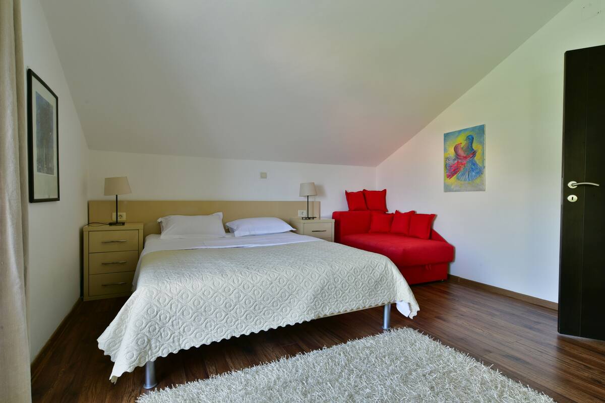 A bedroom with a bed, a red sofa, a white rug, and a painting on the wall.
