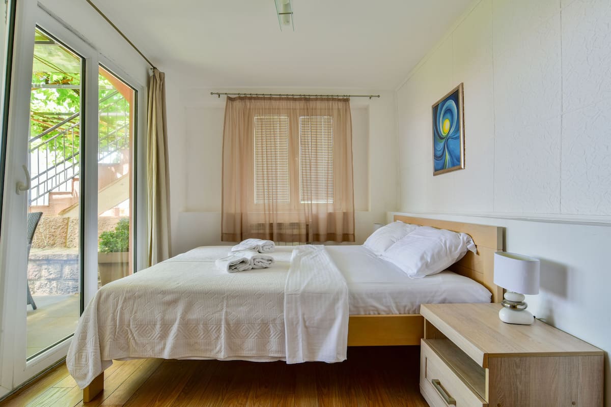 A double bed with a wooden headboard, a bedside table with a lamp, and a window with sheer curtains.