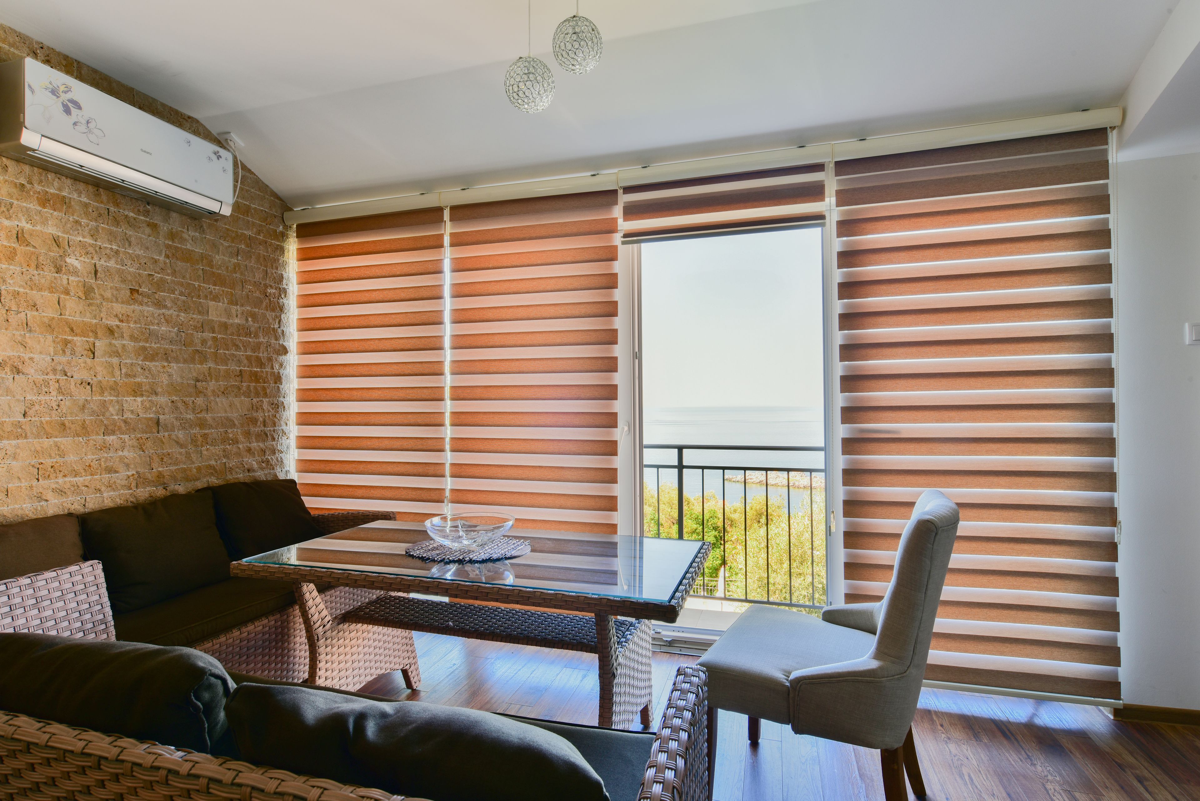 A modern living room with a glass table, wicker furniture, and a view of the outdoors through large windows with blinds.