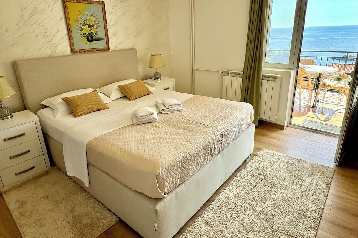A hotel room with a large bed, a nightstand, a lamp, a painting, and a view of the sea through a balcony.