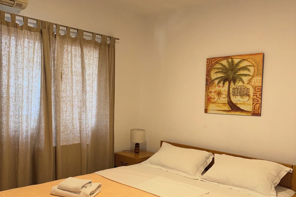 A neatly made bed with white linens and two folded towels, a bedside lamp, and a wall-mounted painting of a palm tree.
