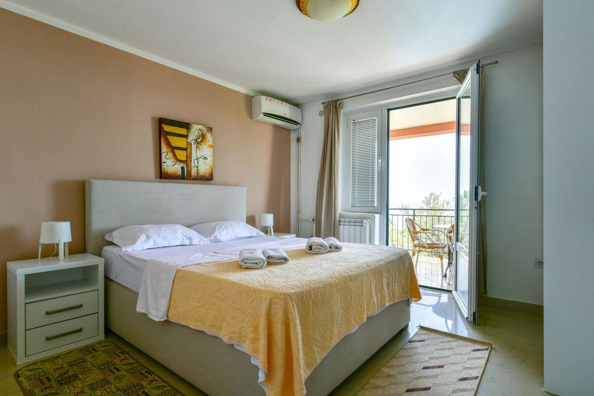 A neatly made bed with white linens and a yellow blanket, a bedside table with a lamp, a painting on the wall, and a balcony with a chair.