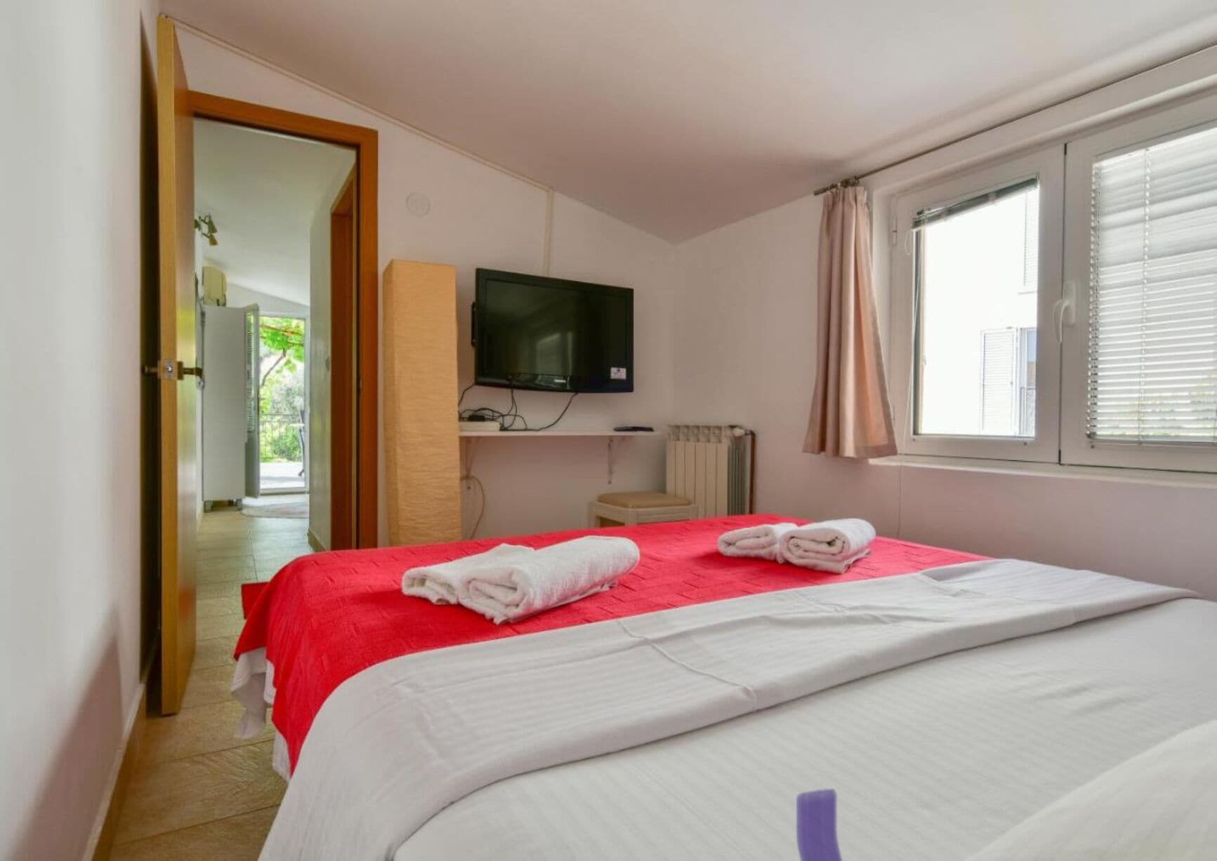 A hotel room with a bed, a TV, a window with blinds, and a bathroom visible through an open door.