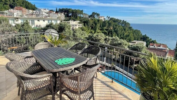 Una terraza con mobiliario de mimbre y piscina, con vistas a un pueblo costero.