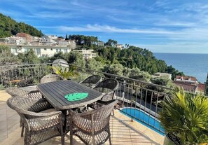 Une terrasse dotée de meubles en osier et d’une piscine, offrant une vue sur une ville côtière.