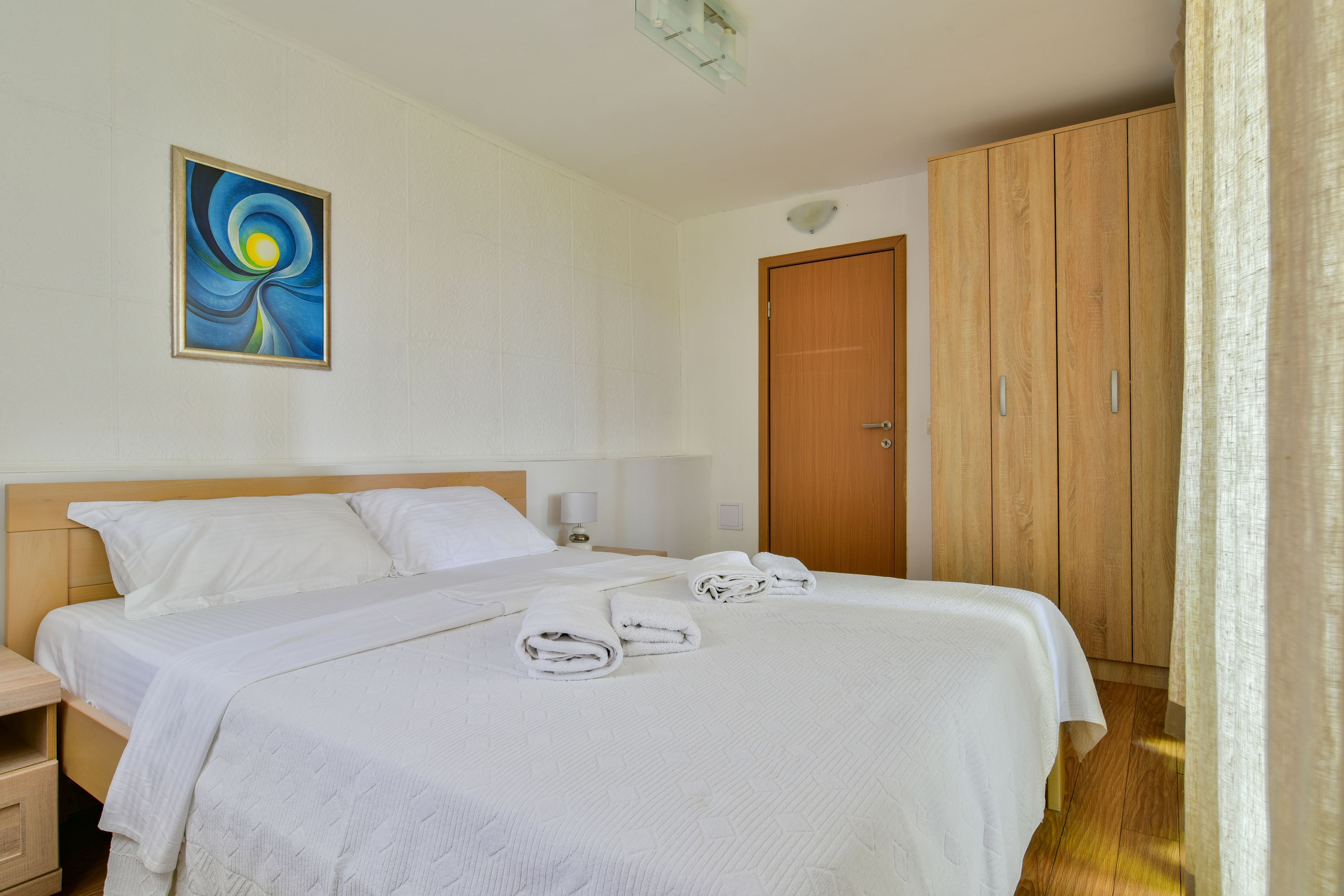 Ein Hotelzimmer mit Bett, Holzschrank, einem Bild an der Wand und einem Nachttisch mit Lampe.
