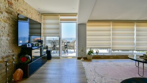 Una sala de estar moderna con una pared de piedra, un televisor de pantalla plana, piso de madera, un ventanal con persianas y un balcón con mobiliario exterior.