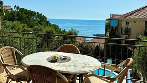Un balcón con sillas de mimbre y una mesa, con vistas a una piscina y al mar.