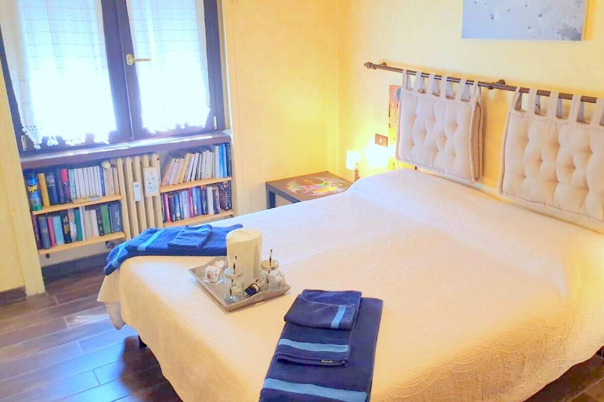 Ein ordentlich bezogenes Bett mit weißer Daunendecke, ein Nachttisch mit Lampe und eine Bücherwand voller Bücher.