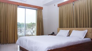 Une chambre d’hôtel avec un grand lit, une tête de lit en bois et une vue sur un balcon à travers des rideaux transparents.