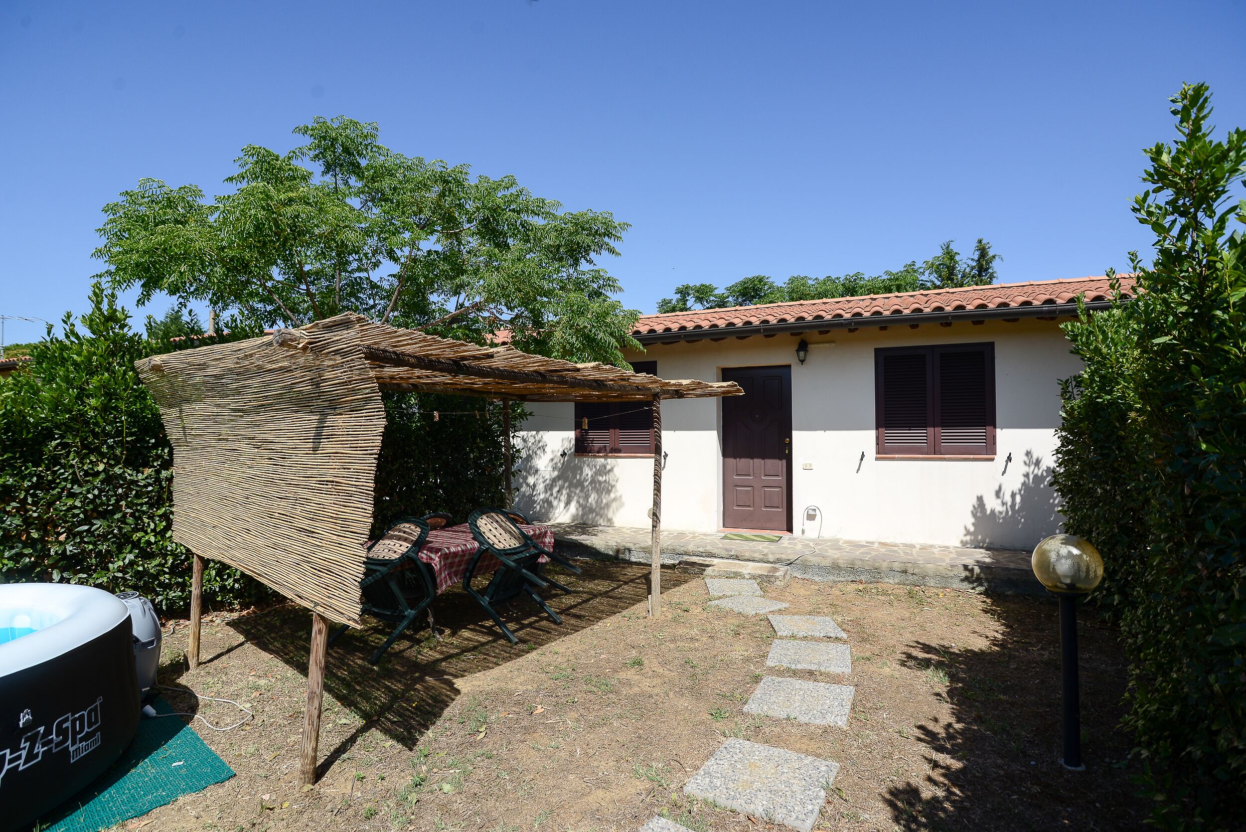 A small house with a tiled roof, a covered patio with a table and chairs, and a swimming pool.