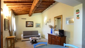 A living room with a wooden ceiling, a sofa, a dining table with chairs, a television, and a cabinet. - Agriturismo Benvenuti (Roccastrada)