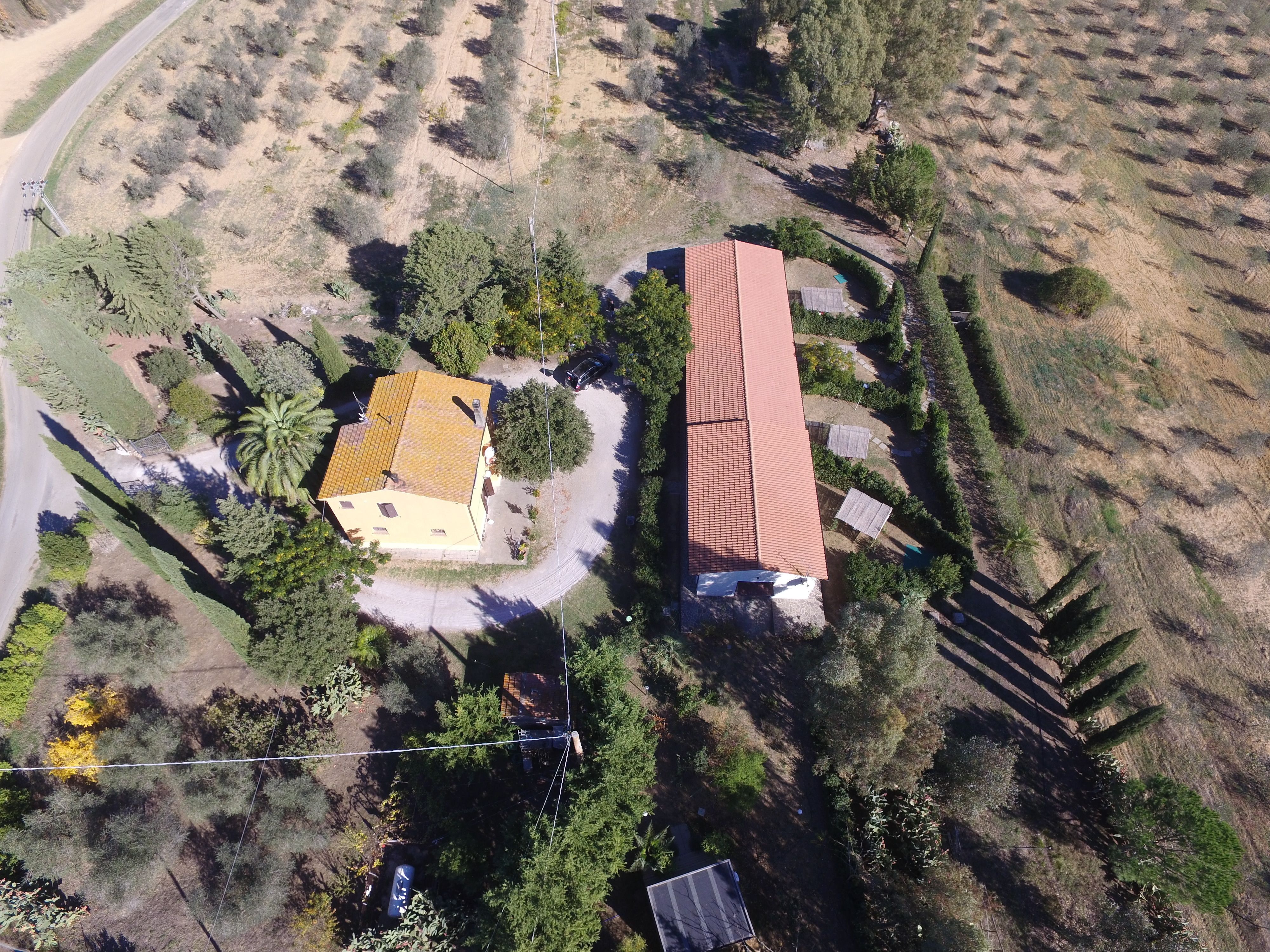 An aerial view of a property with two buildings, one with a yellow facade and another with a red roof, surrounded by trees and a road.