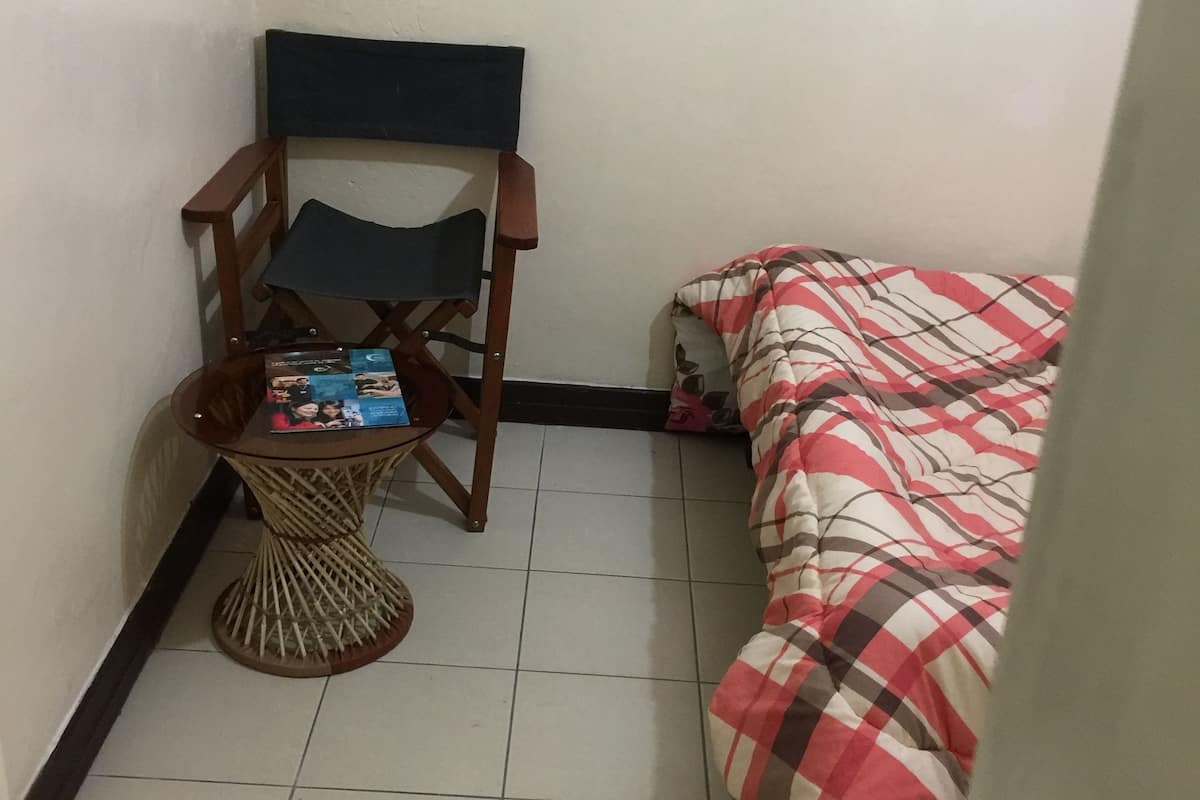 A small, tiled room with a wooden chair, a small table with magazines, and a bed with a checkered blanket.