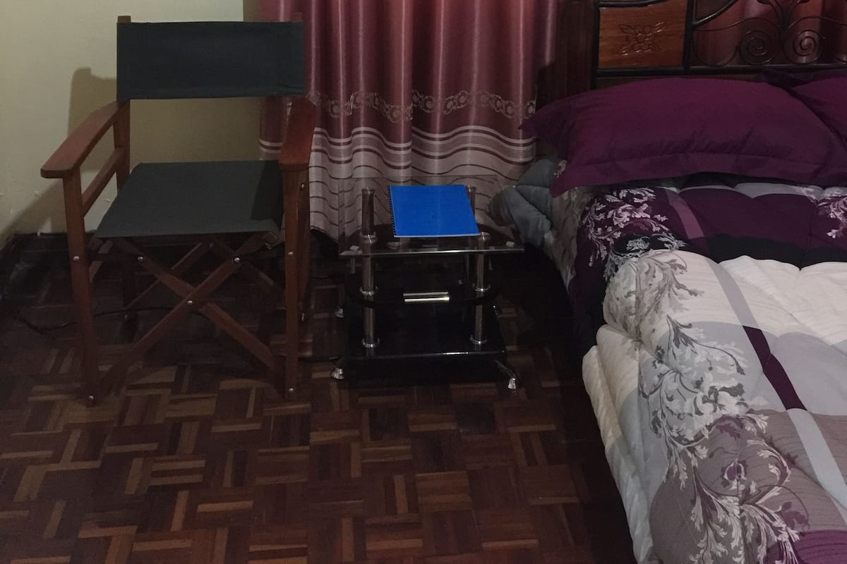 A bedroom with a bed, a chair, a small table with a blue notebook, and a curtain with a floral pattern.