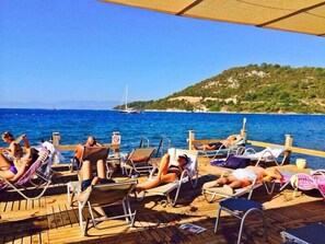 A wooden deck with lounge chairs facing a body of water, a sailboat in the distance, and a hilly landscape in the background. - Gunisigi Otel (Bodrum)
