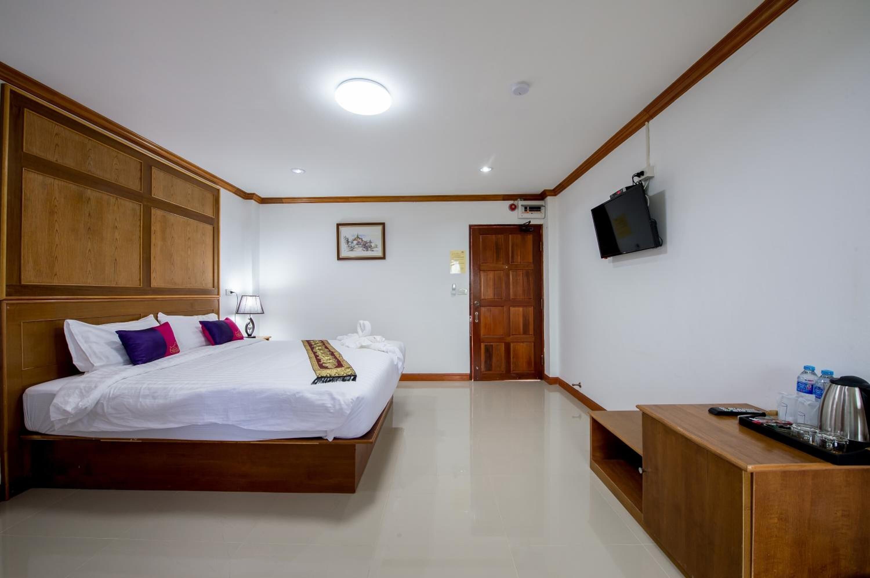 A hotel room with a wooden bed, a TV mounted on the wall, a wooden desk with a kettle, and a door leading to another room.