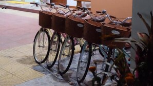 Een rij fietsen met mandjes, geparkeerd op een betegelde vloer in een openbare ruimte met een houten deur en een glazen scheidingswand op de achtergrond.