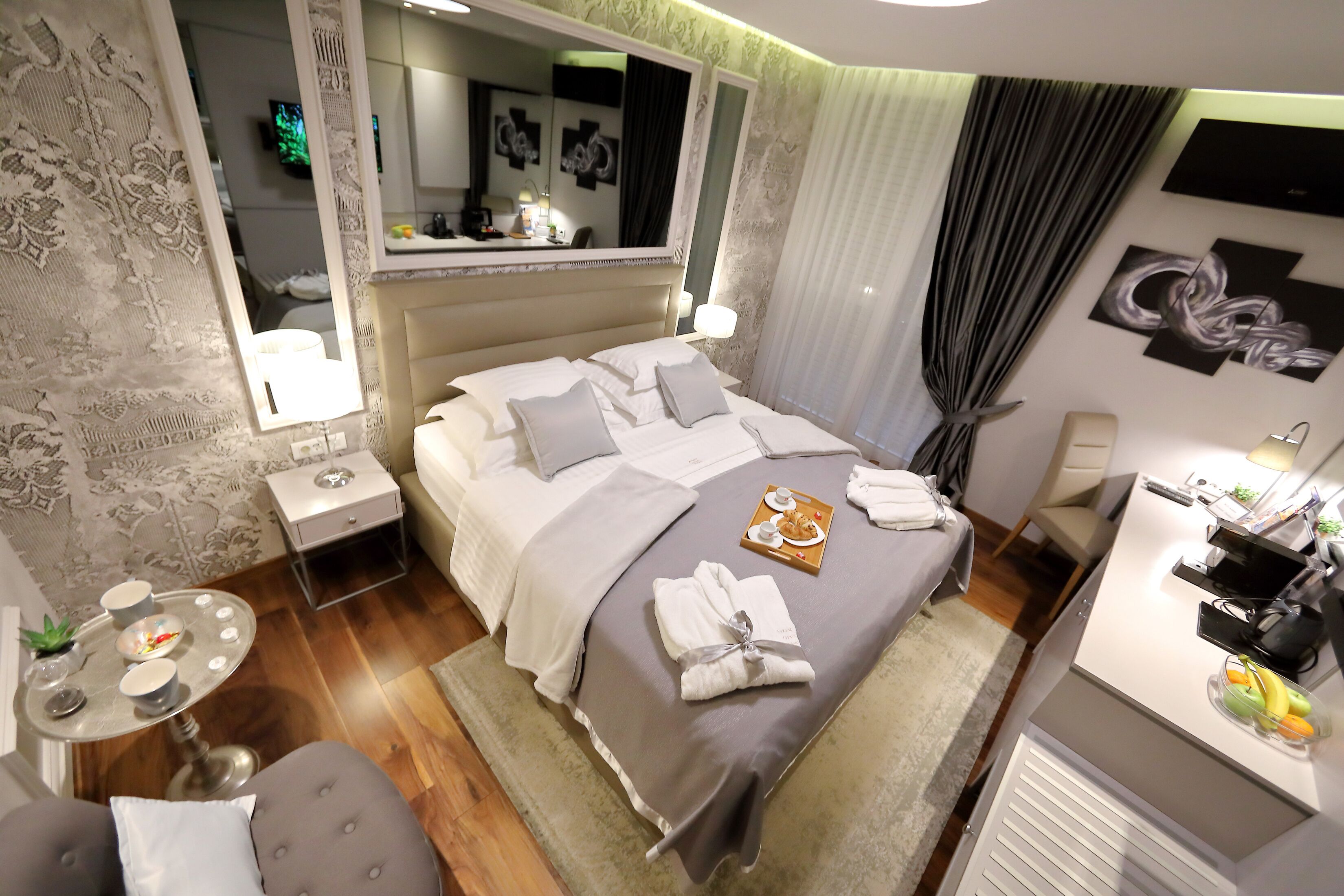 A hotel room with a large bed, a bedside table, a small round table, a chair, a desk with a chair, a mirror, and artwork on the walls.
