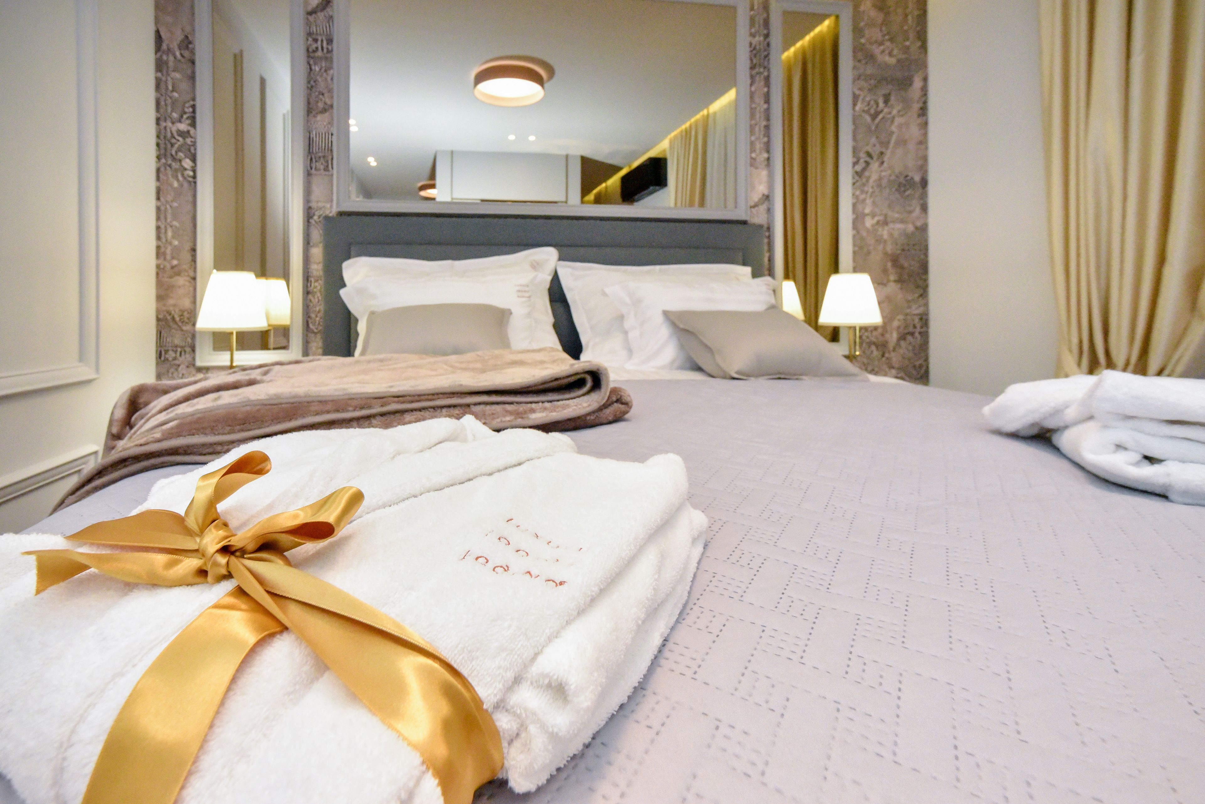 A neatly made bed with white linens and a gold ribbon, a headboard, two bedside lamps, and a mirror above the bed.