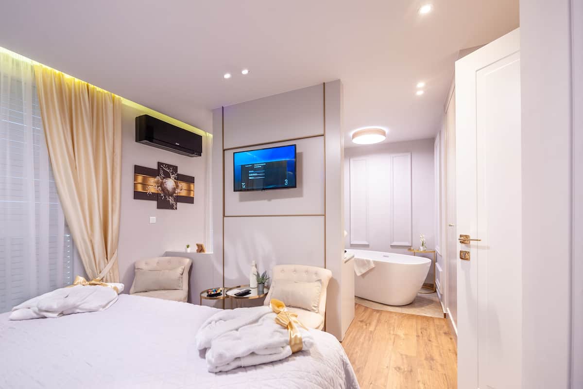 A modern hotel room with a large bed, a flat-screen TV, a bathtub, and a small side table with a remote control.