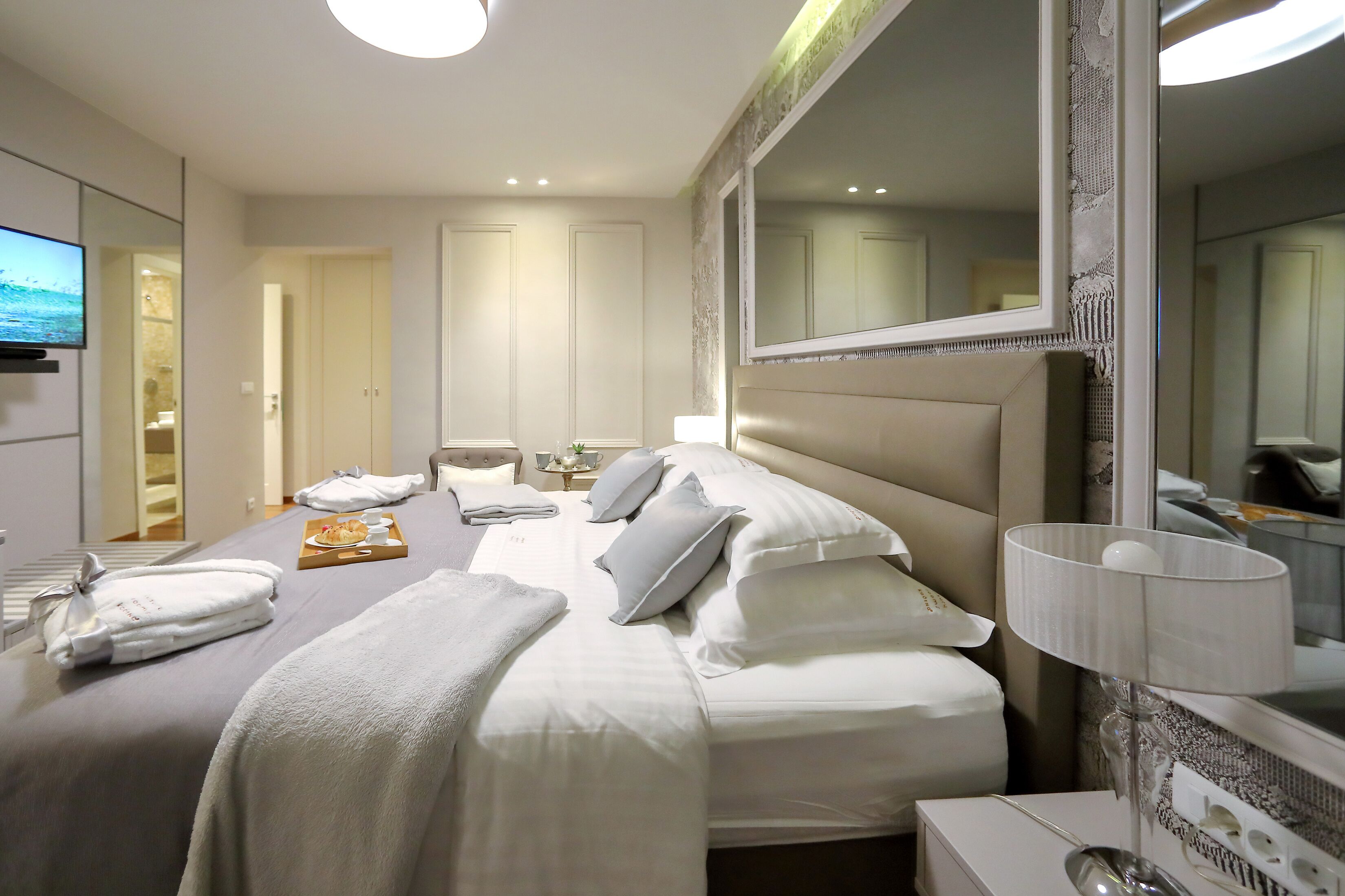 A hotel room with a large bed, a bedside table with a lamp, a TV, and a bathroom visible through an open door.