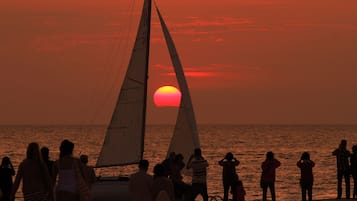 Um pôr do sol com um veleiro e pessoas na praia.