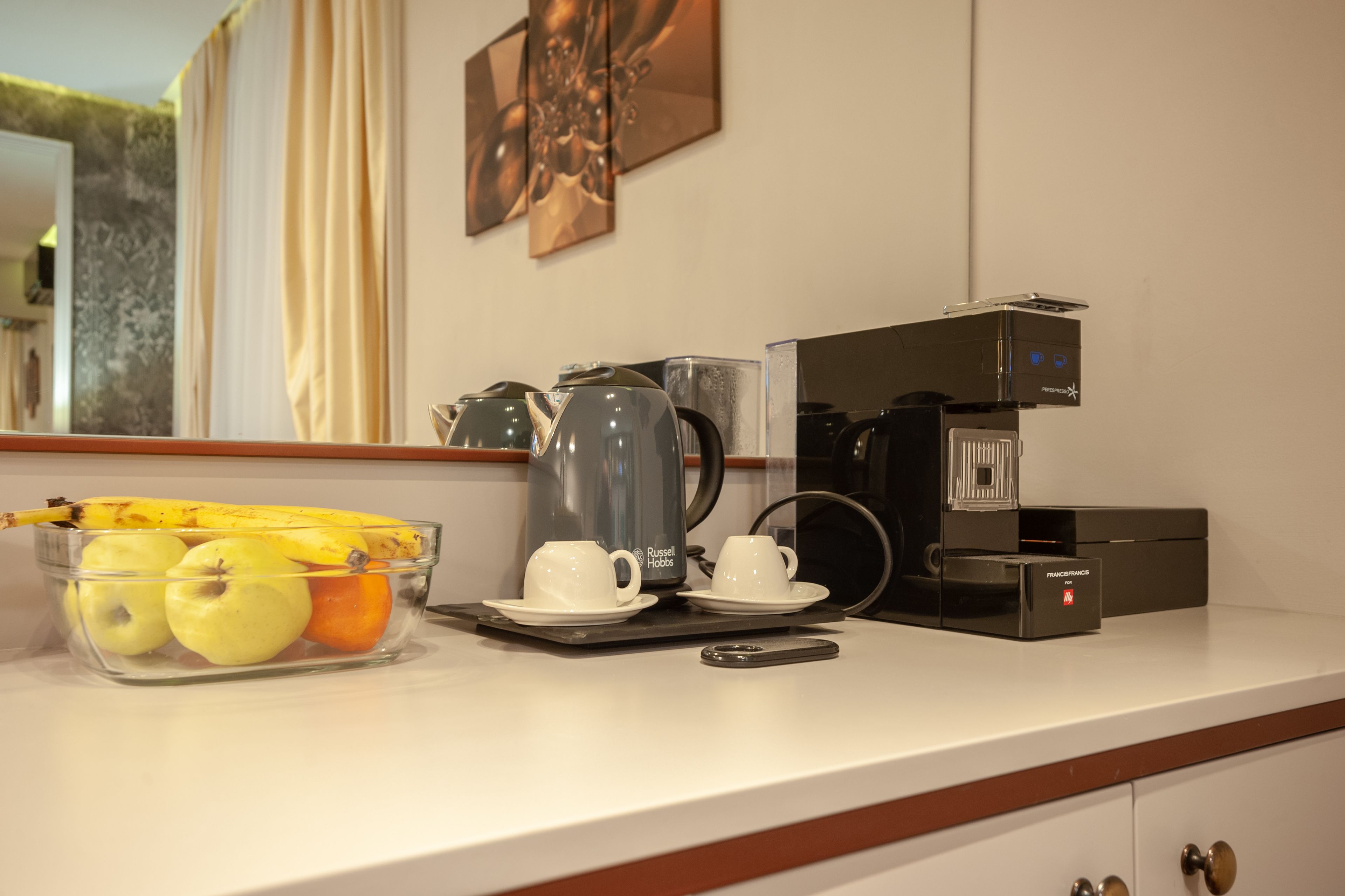A kitchen counter with a coffee maker, two cups, a kettle, and a fruit bowl containing apples and a banana.
