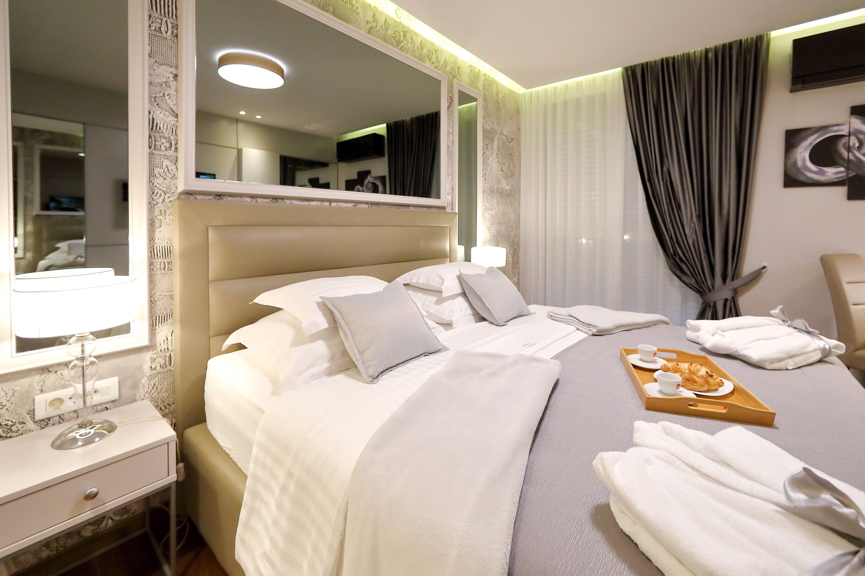 A neatly made hotel room with a large bed, a bedside table with a lamp, a tray with breakfast items, and a large mirror.