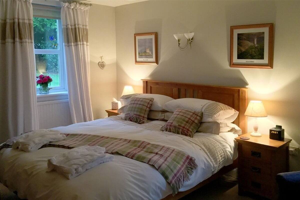 A neatly made bed with pillows and a blanket, bedside tables with lamps, a window with curtains, and wall-mounted pictures.