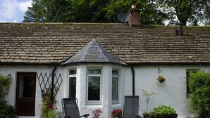 A white cottage with a tiled roof, two outdoor chairs, and a garden with potted plants. - Druminoch (Kirriemuir)