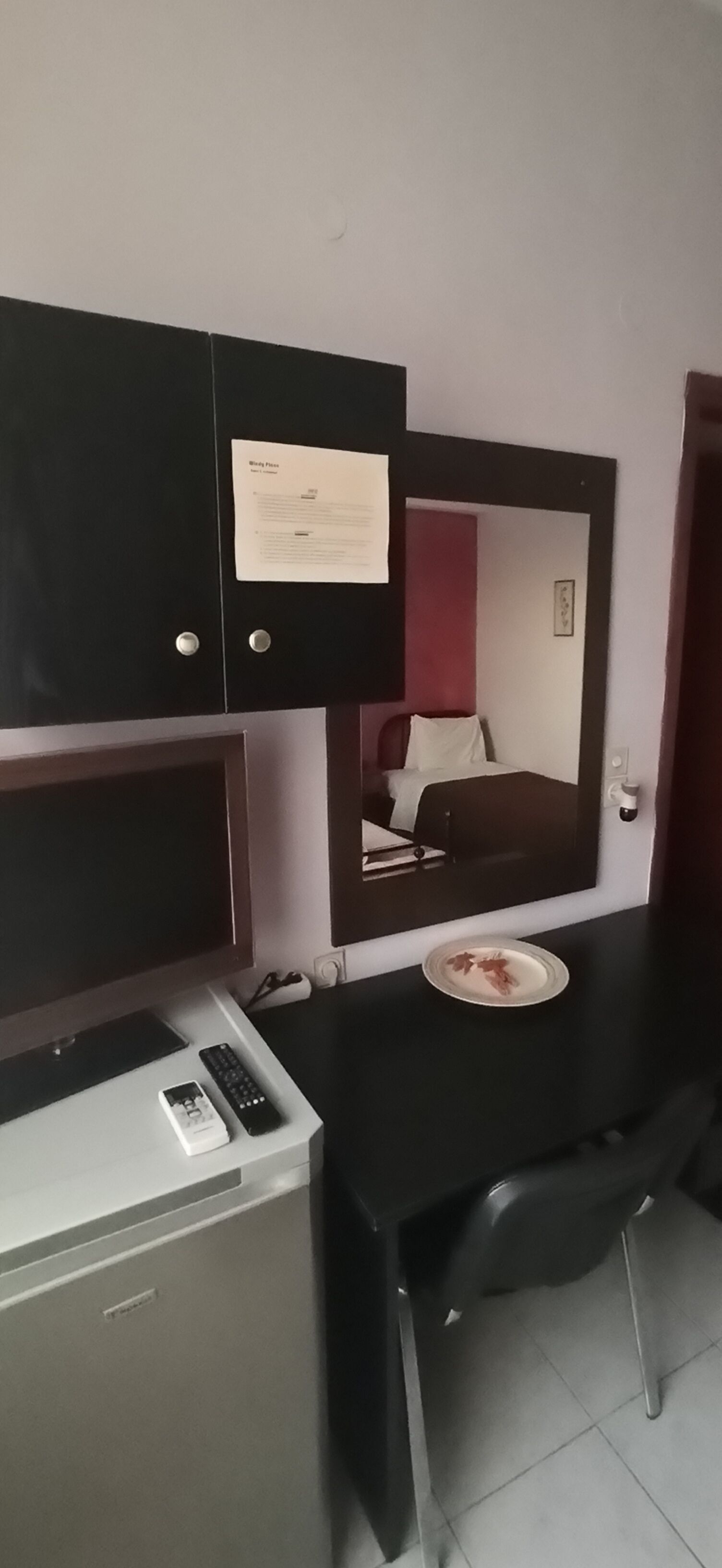 Habitación de hotel compacta con cama, escritorio, espejo, refrigerador pequeño y mesita de noche con control remoto y teléfono.