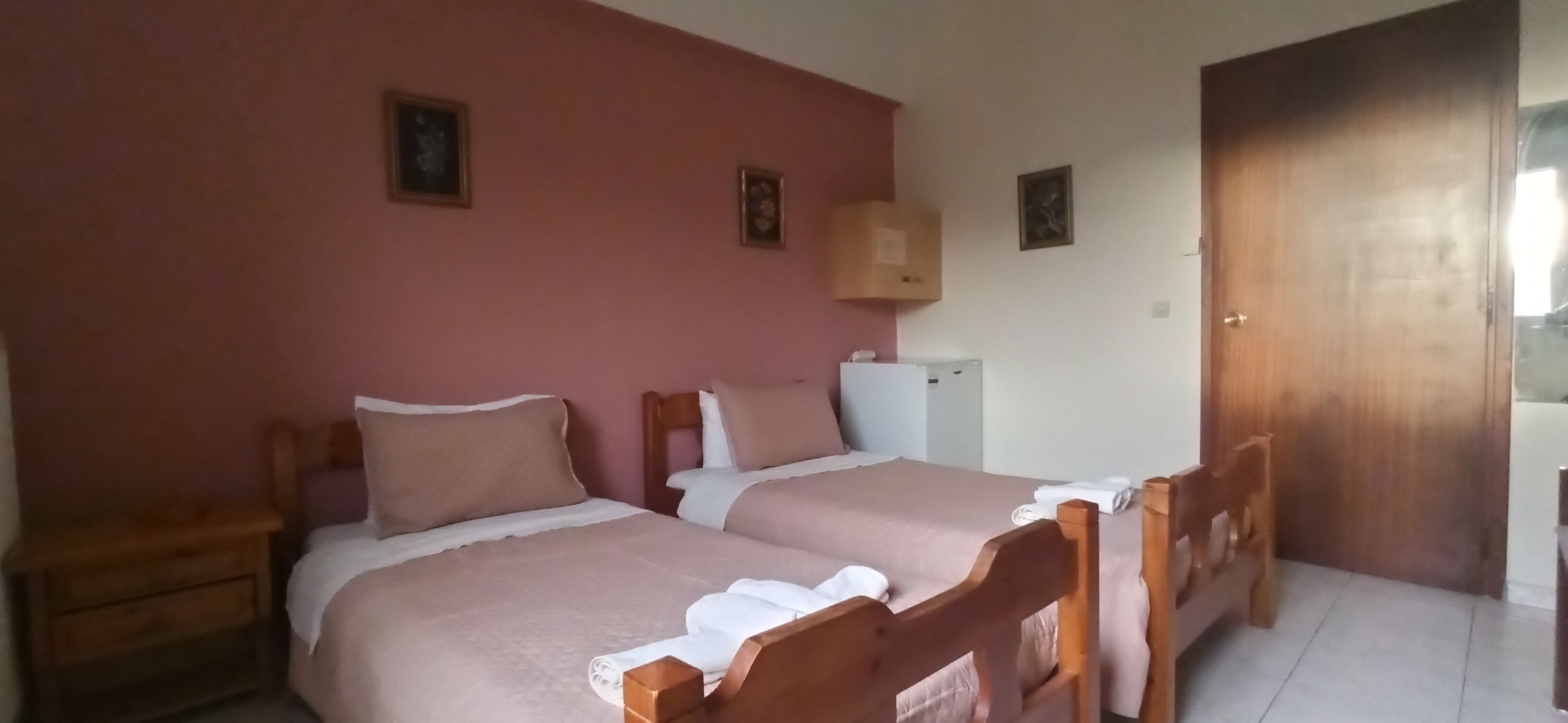 A room with two beds, wooden headboards, a small white refrigerator, a wooden cabinet, and a door.