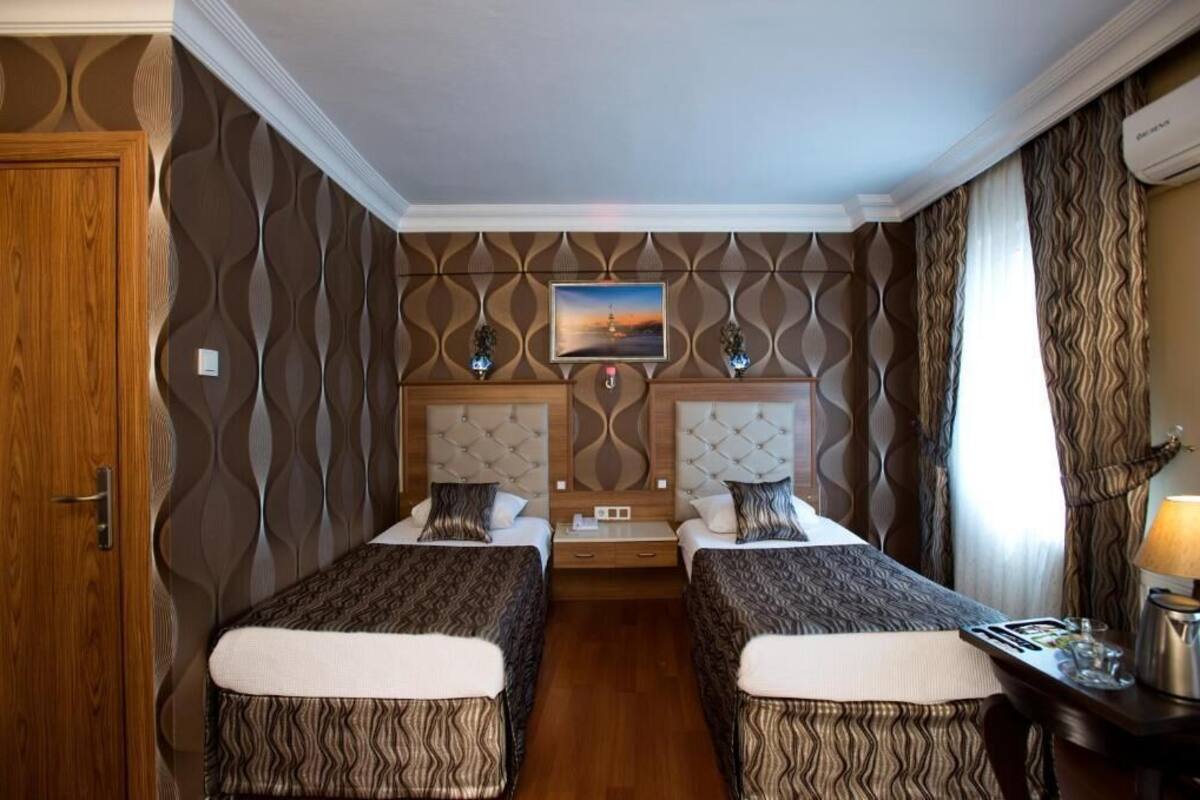 A hotel room with two beds, a wooden door, patterned wallpaper, a painting on the wall, and a nightstand with a lamp.