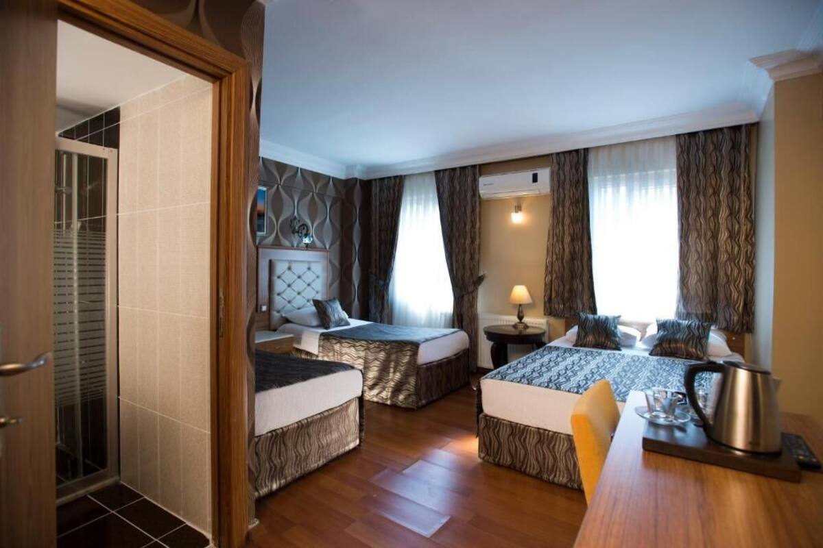A hotel room with two beds, a wooden floor, a small table with a kettle, and a window with curtains.