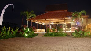 Un espacio al aire libre cubierto para comer, con mesas y sillas, rodeado de plantas tropicales y un sendero pavimentado.