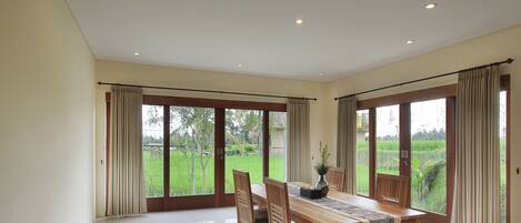 A spacious dining area with a wooden table and chairs, large windows with curtains, and a view of a green landscape.