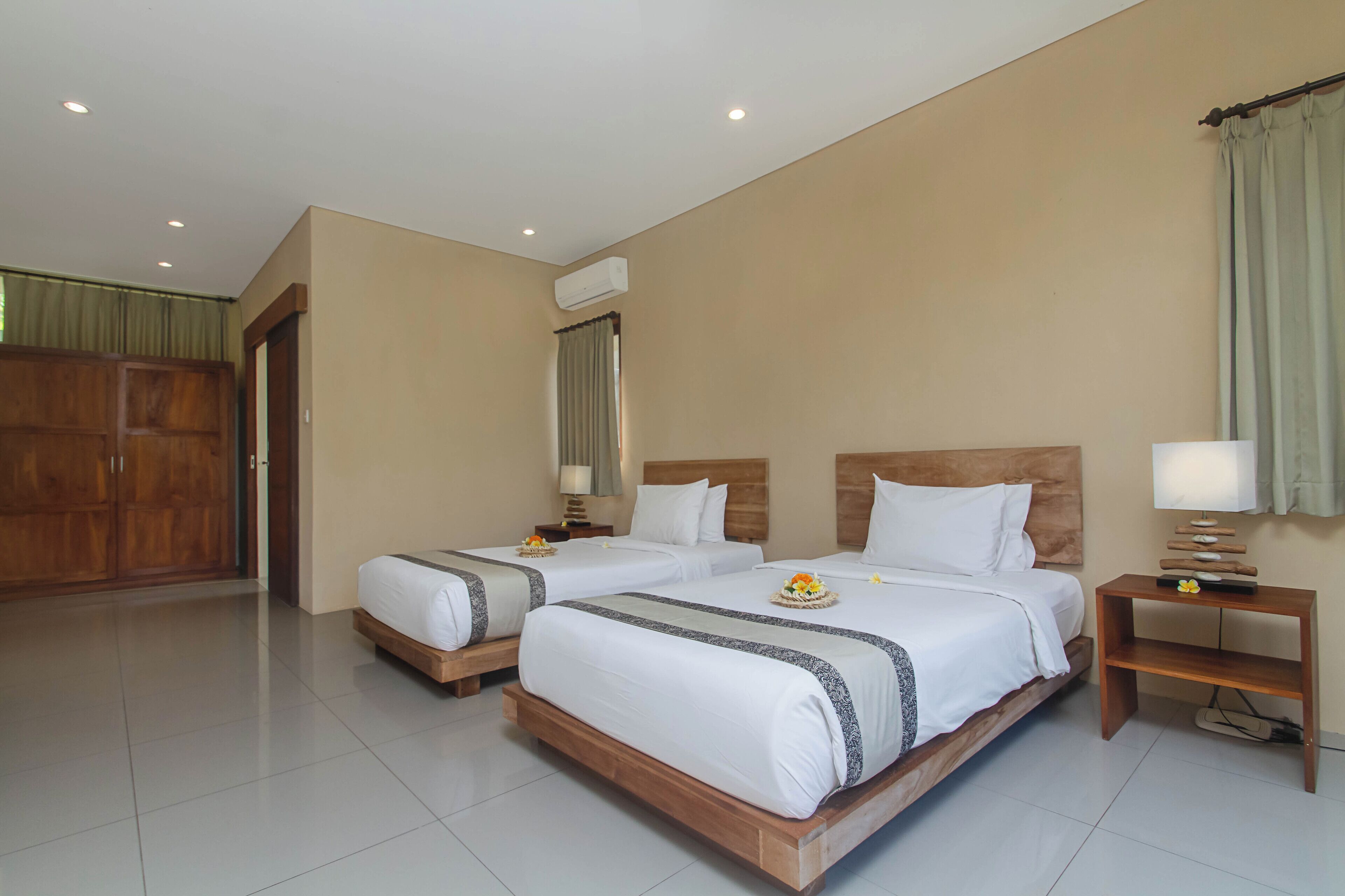 A hotel room with two beds, a wooden wardrobe, an air conditioning unit, and a nightstand with a lamp.